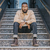 A person with glasses and a beard sits on a stairway, wearing a beige jacket, rolled-up jeans, and brown boots, with hands resting on knees.