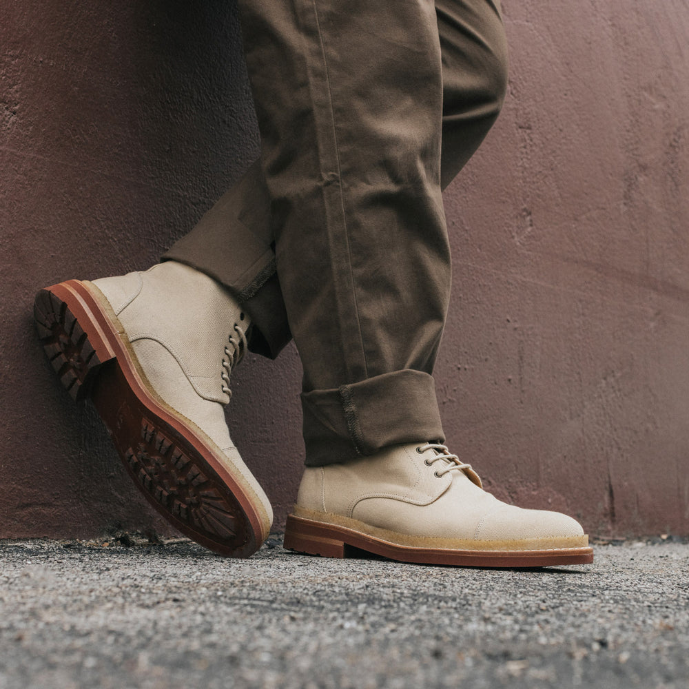 Person wearing beige boots and brown pants, standing against a brick wall with one leg crossed over the other.