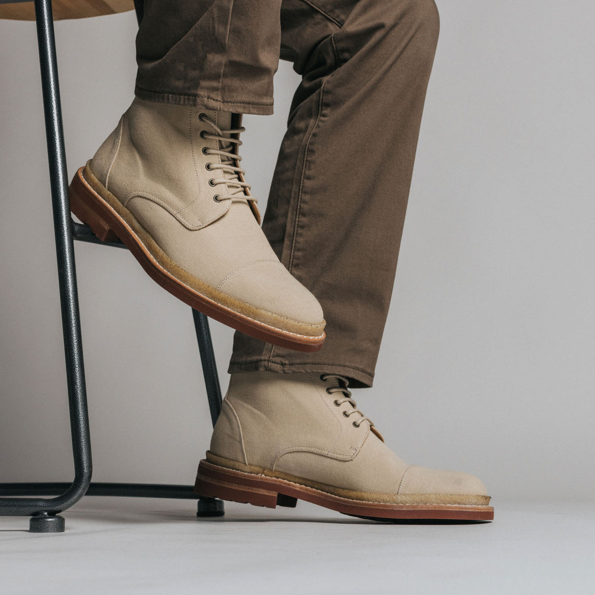 A person wearing tan lace-up boots and brown pants sits on a high stool, with their legs crossed and the lower part of their body visible.