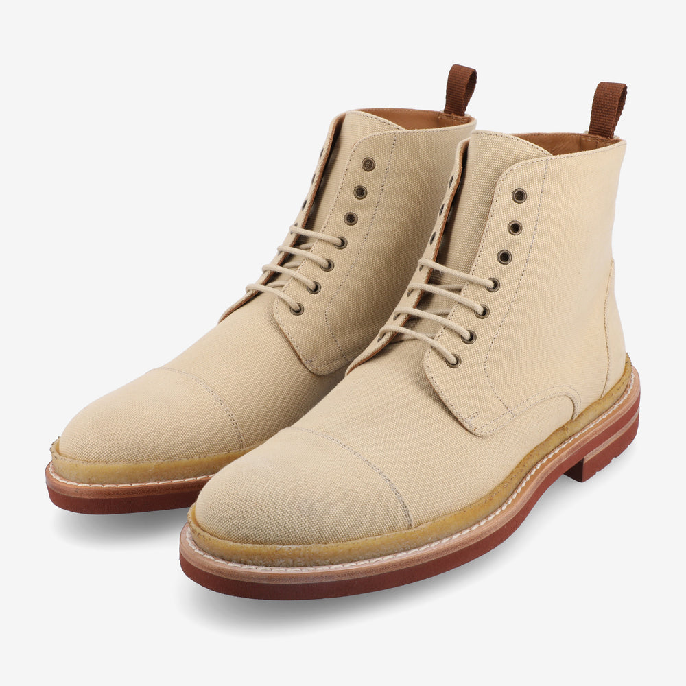 A pair of beige canvas boots with brown soles and laces, featuring a pull tab at the back and metal eyelets for the laces.