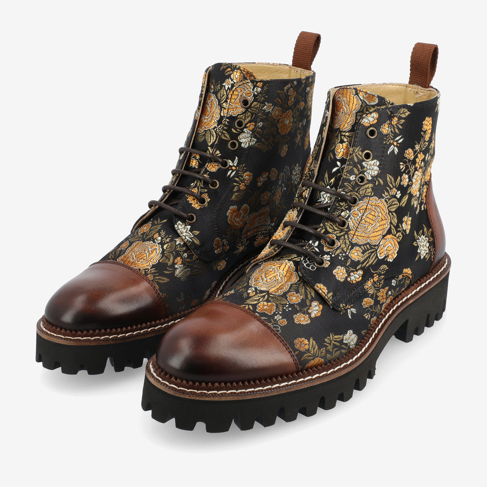 A pair of high-top lace-up boots with a floral pattern, featuring brown leather toe caps and heels, and rugged black soles.