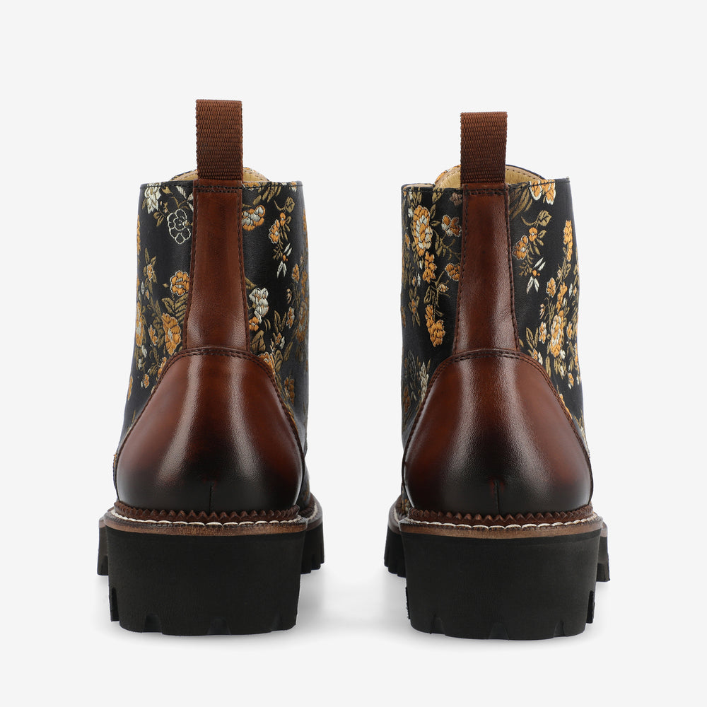 Back view of a pair of ankle boots with brown leather heels and black fabric uppers featuring a floral pattern. The boots have pull tabs and black lugged soles.