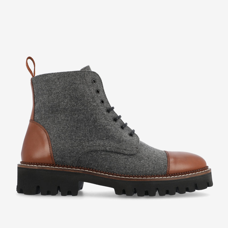 A grey high-top boot with brown leather accents on the toe and heel, featuring black laces, black lug sole, and a pull loop at the back.