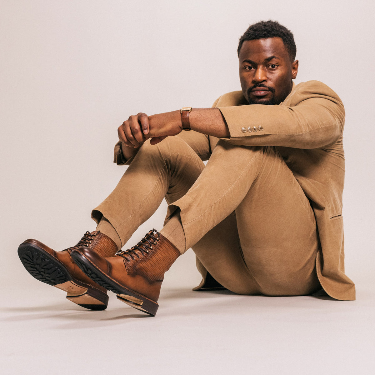 A man in a tan suit and brown boots sits on the ground with his arms resting on his knees, looking forward.