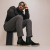 A man in a black shirt and grey pants sits on a black cube, resting his head on his hand, looking to the side. He wears black suede chelsea boots.