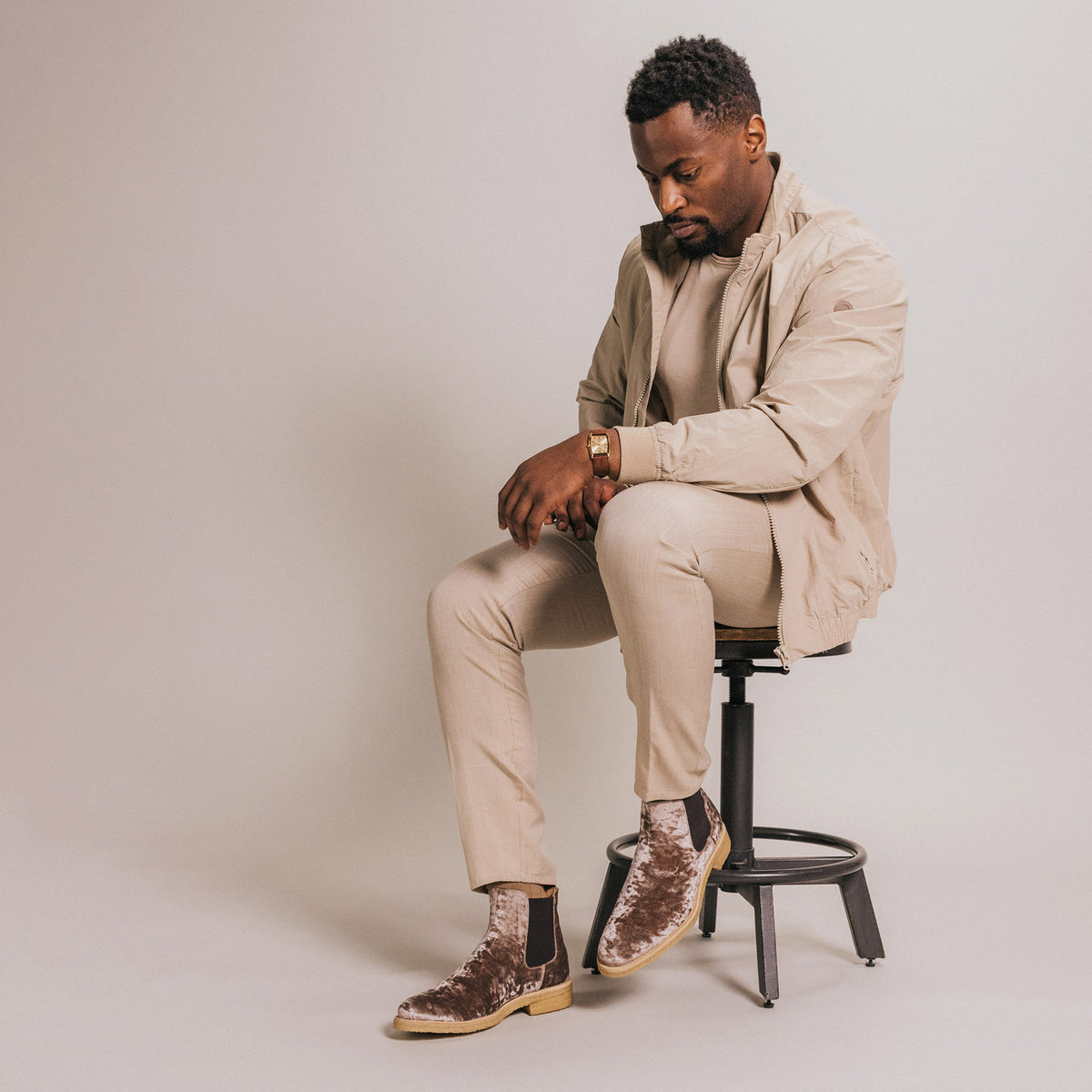 A man sits on a stool wearing a light beige outfit with matching jacket and pants, complemented by patterned velvet boots. He looks down while adjusting his sleeve.