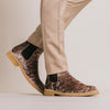 A person wearing beige pants and brown velvet Chelsea boots with light tan soles stands against a neutral background.