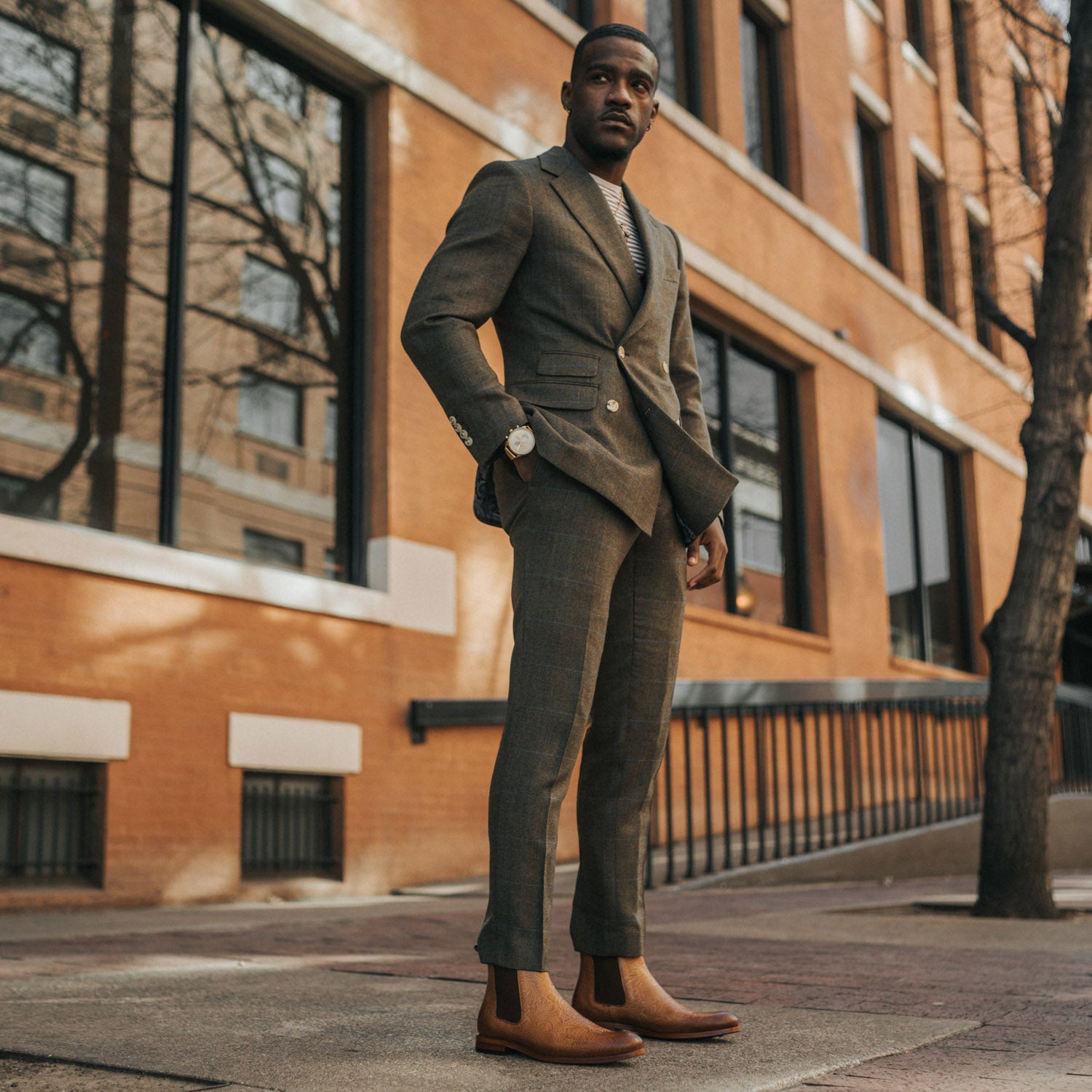 A man stands confidently on a city sidewalk wearing a brown suit, striped shirt, and brown boots, with a brick building in the background.