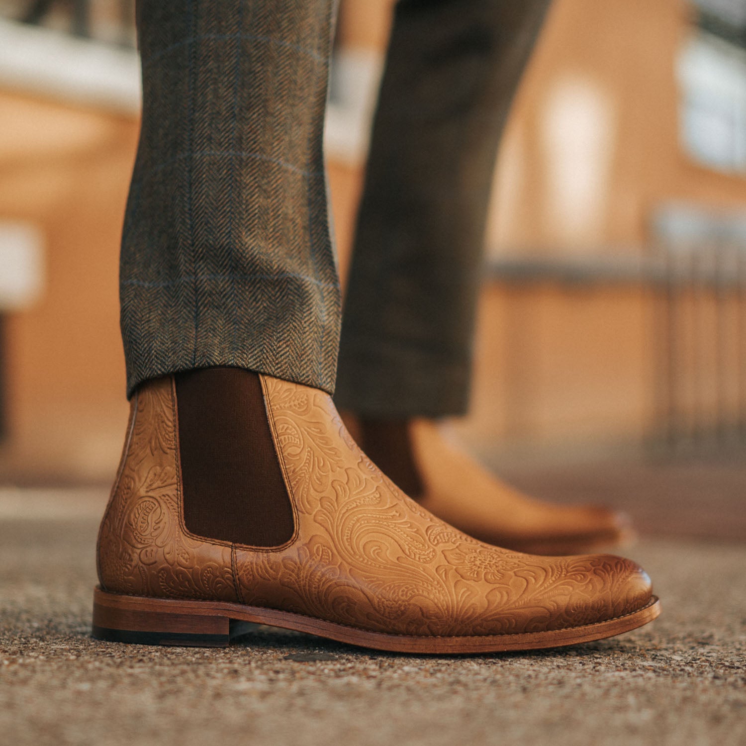 A close-up of a person wearing brown, intricately patterned leather Chelsea boots paired with dark tweed pants.