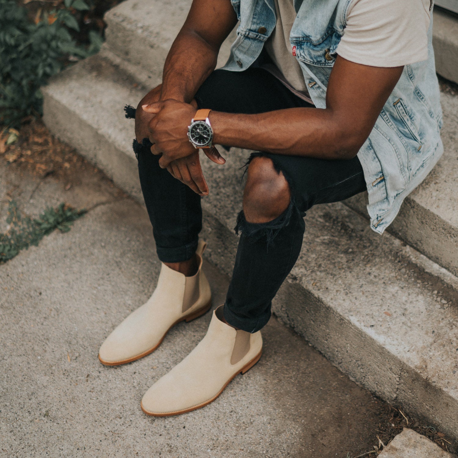 Person sitting on concrete steps wearing a sleeveless denim jacket, white shirt, ripped black jeans, beige boots, and a watch.