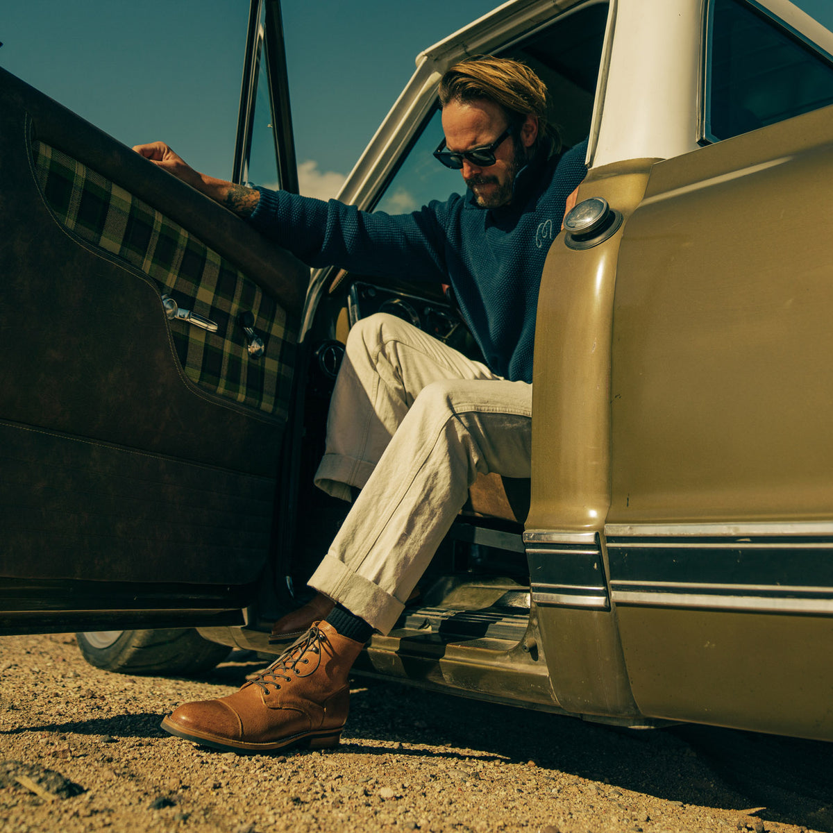 A person wearing sunglasses and casual clothes steps out of an open car door, partially seated inside a vintage vehicle.