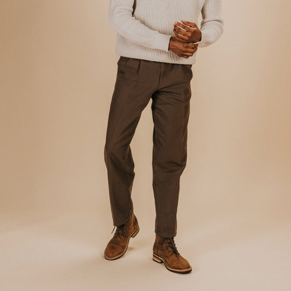 Person wearing a light gray sweater, brown pants, and brown boots, standing against a plain beige background. The person's face is not visible.