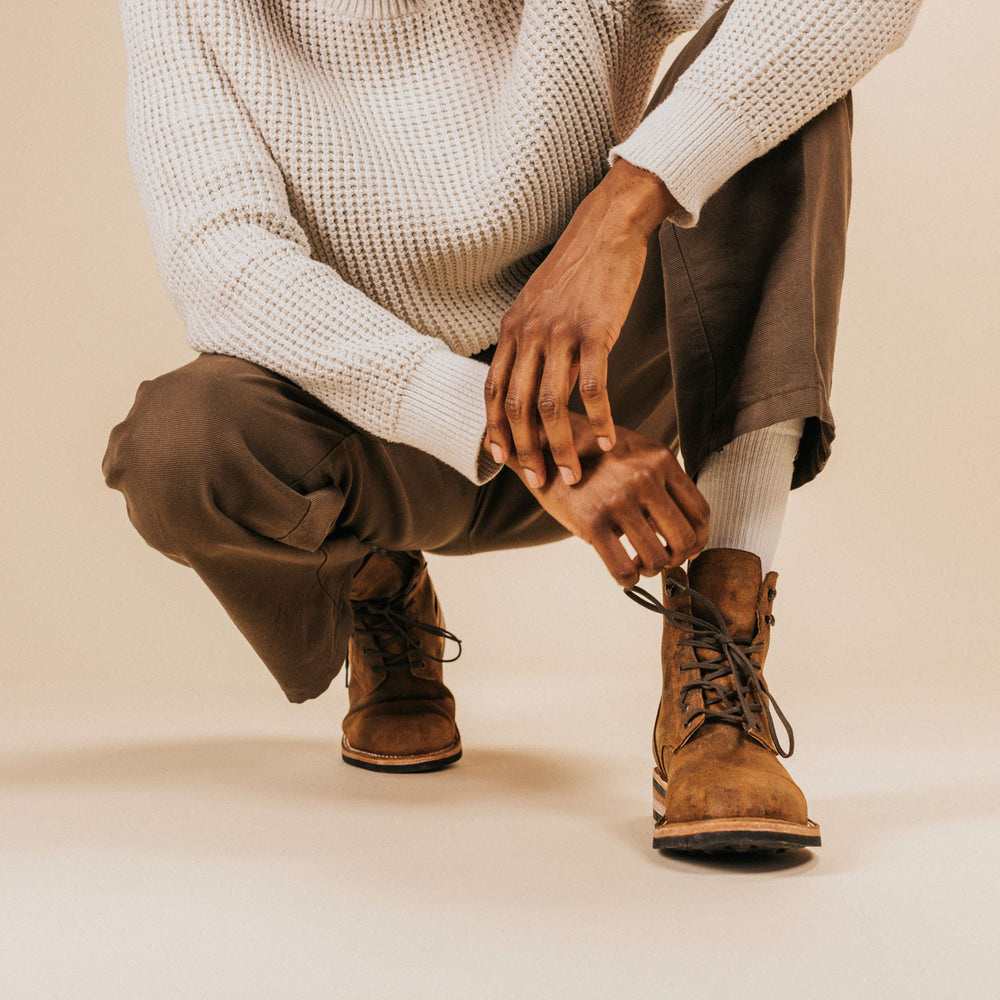 Person wearing a white sweater, brown pants, and brown boots, crouching down, with one hand resting on their knee and the other hand touching the ground.