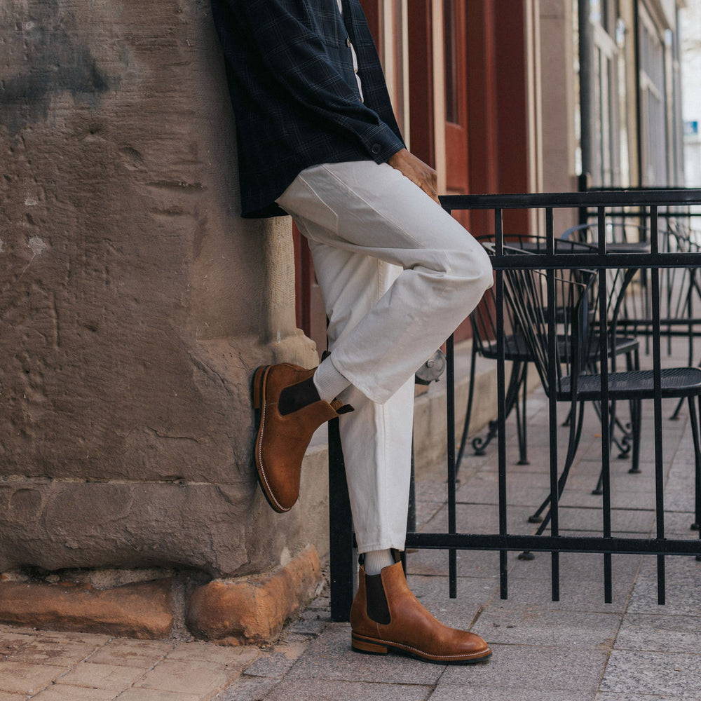 A person leans against a stone wall in white pants and brown boots, beside an empty outdoor caf√© table and chairs.