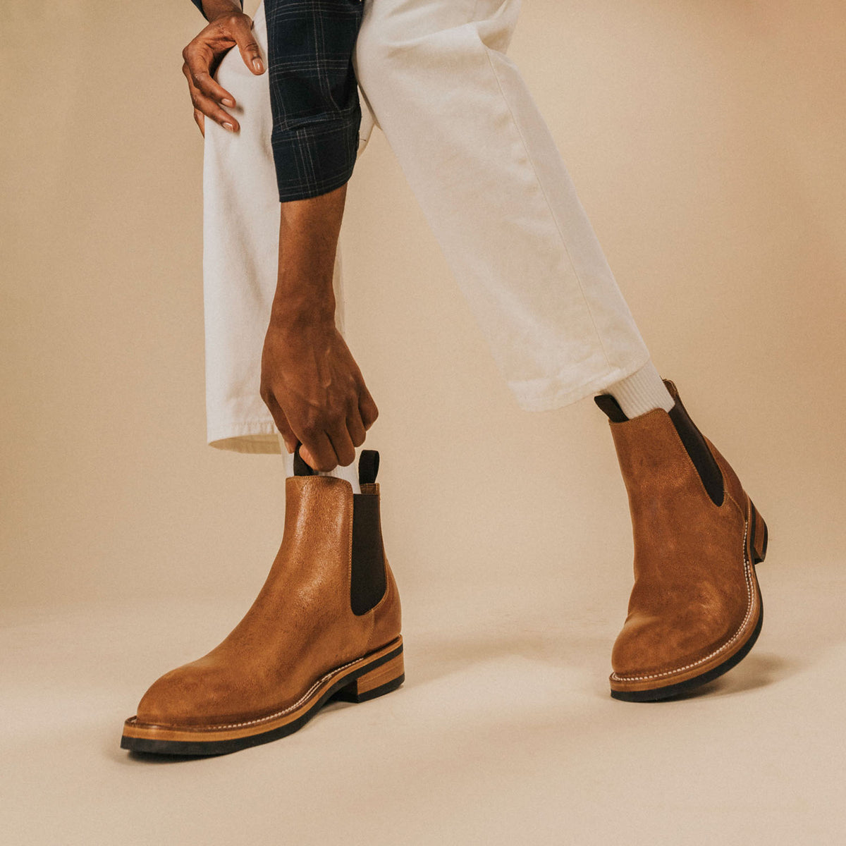 A person tucking their pant leg into a light brown Chelsea boot on their foot, while wearing white pants.