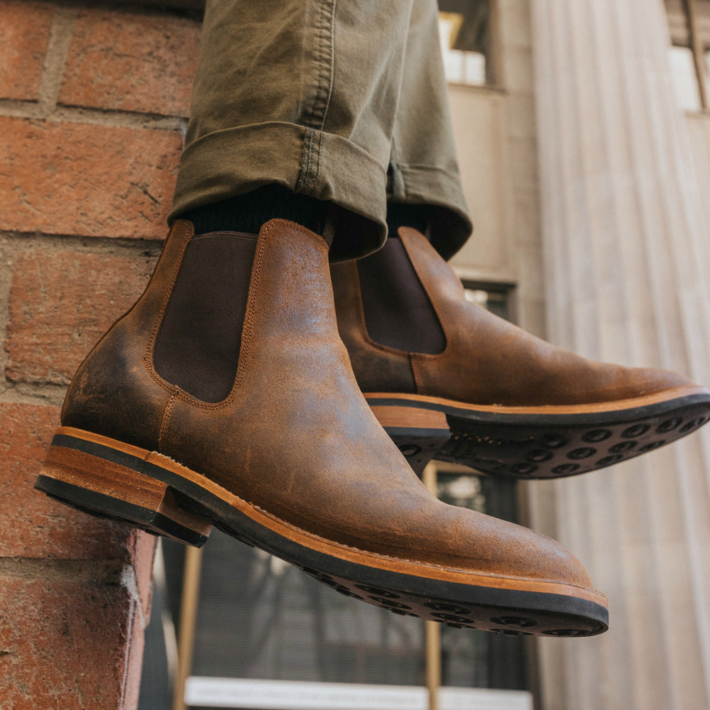 Close-up of a pair of brown leather Chelsea boots worn with olive green pants. The person is seated with their legs extended, showing the boots' details against a brick wall and building background.