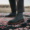 A person wearing gray wool and black leather lace-up boots stands on a patterned rug outdoors.