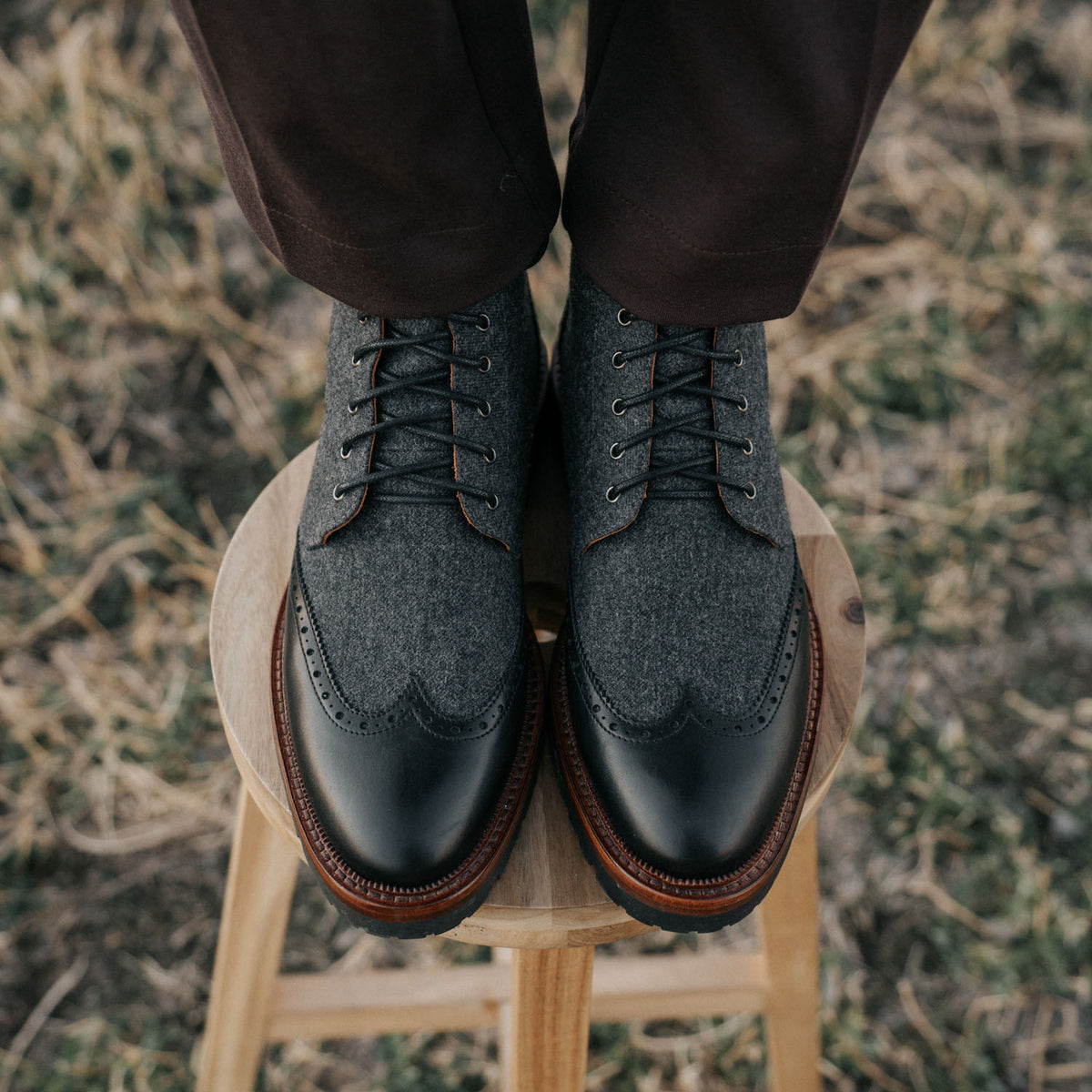 Close-up of a person standing on a wooden stool wearing gray and black leather wingtip shoes and brown pants. The background features a grassy, outdoor setting.