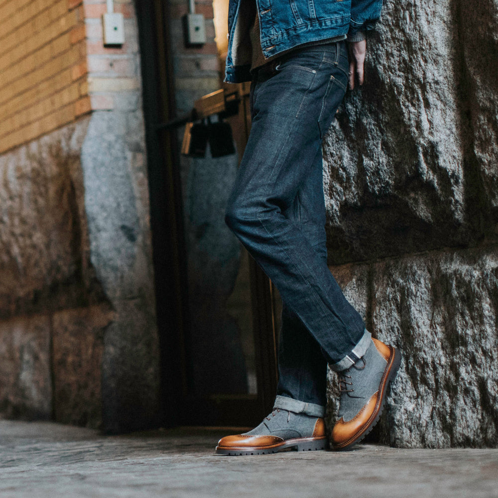 A person in a denim jacket, rolled-up jeans, and brown leather shoes stands with their legs crossed, leaning against a stone wall in an urban setting.