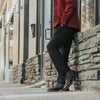 Person in a red jacket and black pants leaning against a stone wall alongside a sidewalk. Only the lower half of the body is visible.