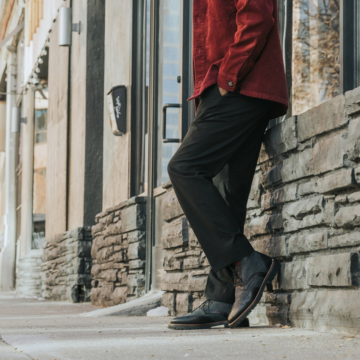 Person in a red jacket and black pants leaning against a stone wall alongside a sidewalk. Only the lower half of the body is visible.