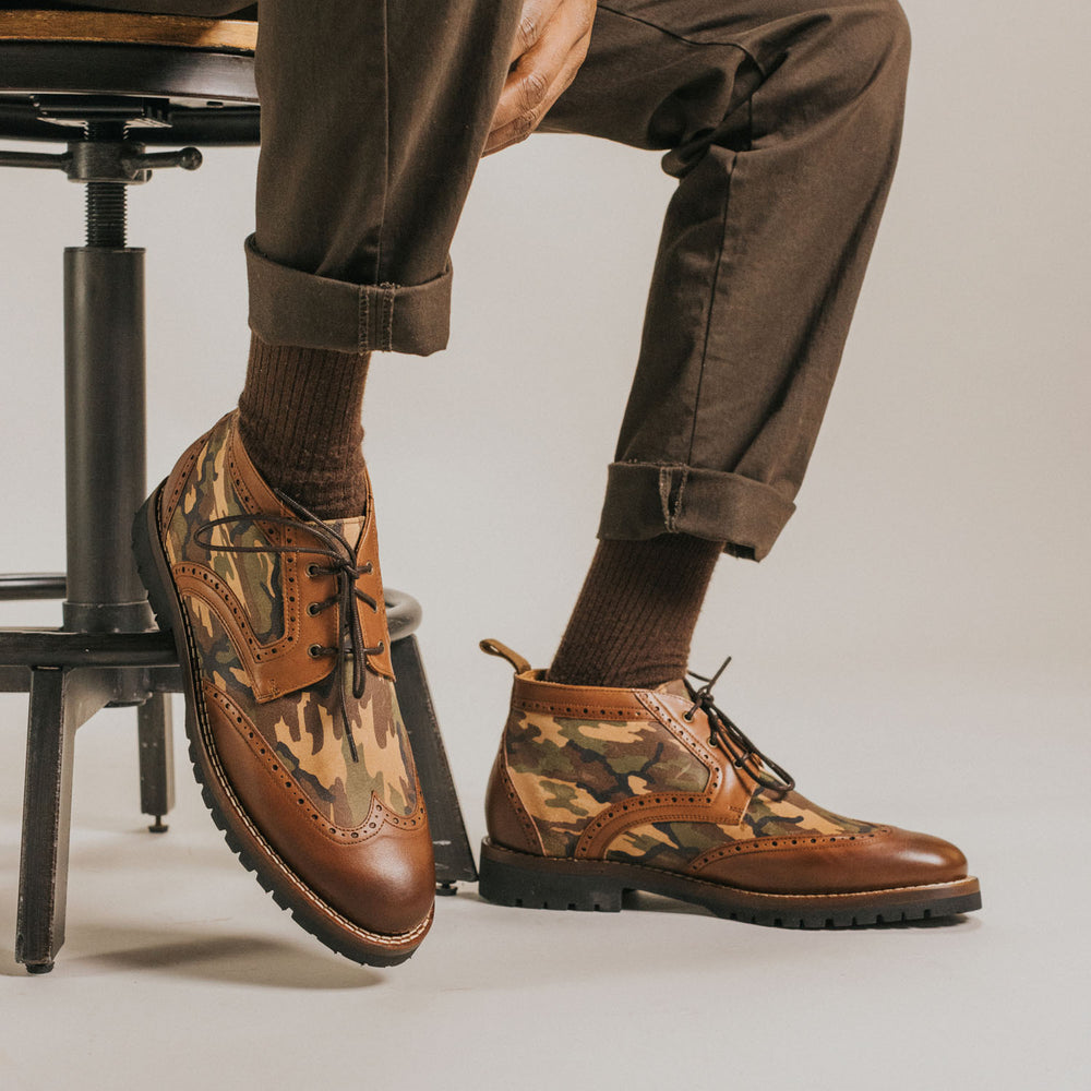 Person seated on a stool wearing brown trousers rolled at the cuffs, brown socks, and camouflage-patterned lace-up boots with brown trim.