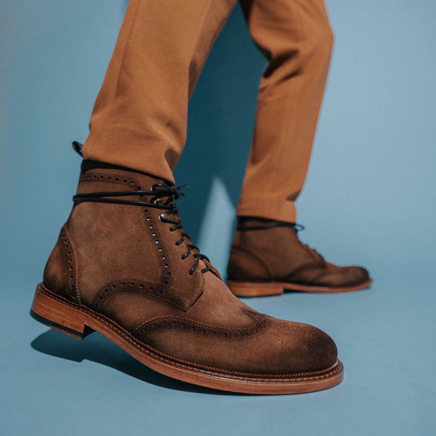 Close-up of a person's legs wearing brown dress boots and tan pants, with a blue background.