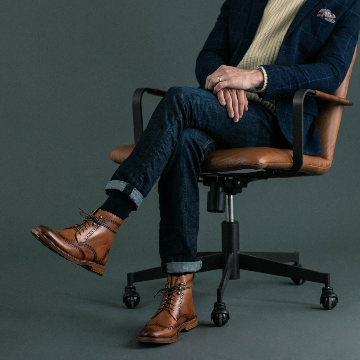 Person seated in an office chair wearing rolled-up jeans, a navy blazer, a cream sweater, and brown leather boots with black socks.