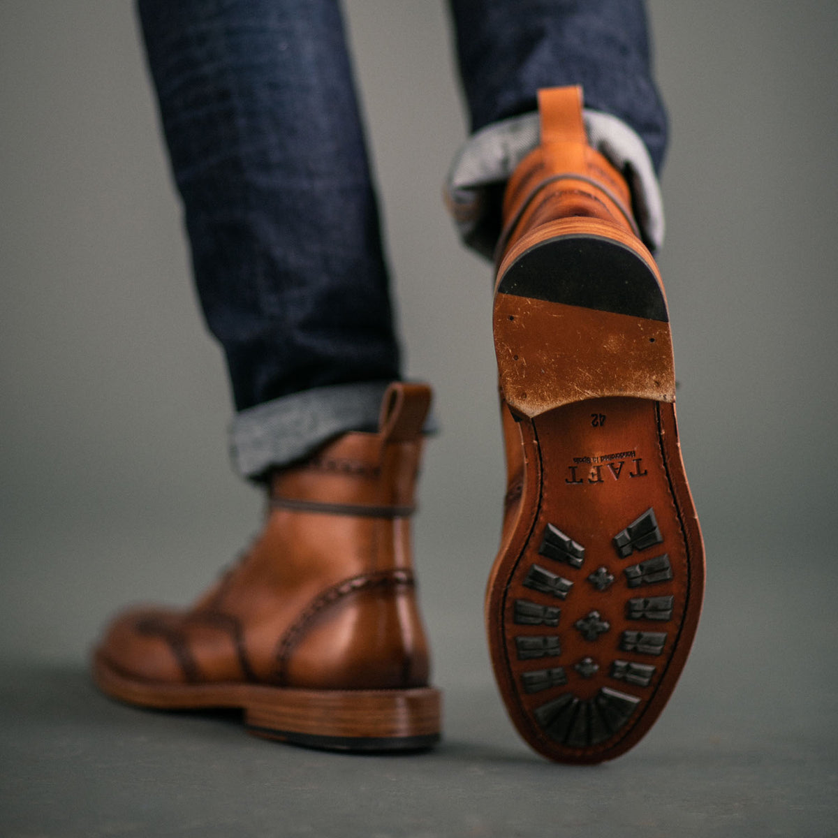 Close-up of a person wearing brown leather boots with visible sole branding and a rolled-up hem on blue jeans.