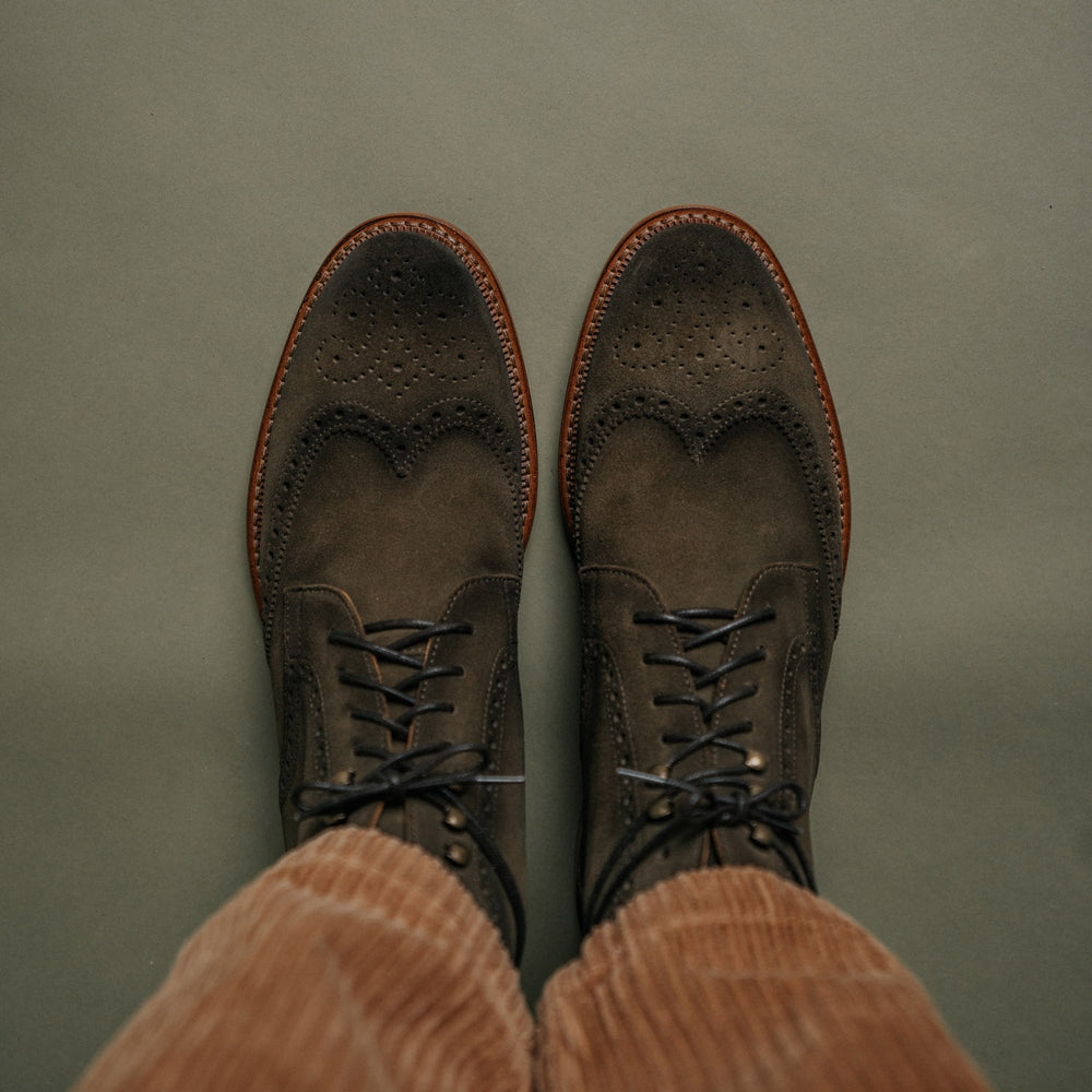 A top view of a person wearing brown suede lace-up dress shoes with perforated detailing and light brown corduroy pants against a green background.