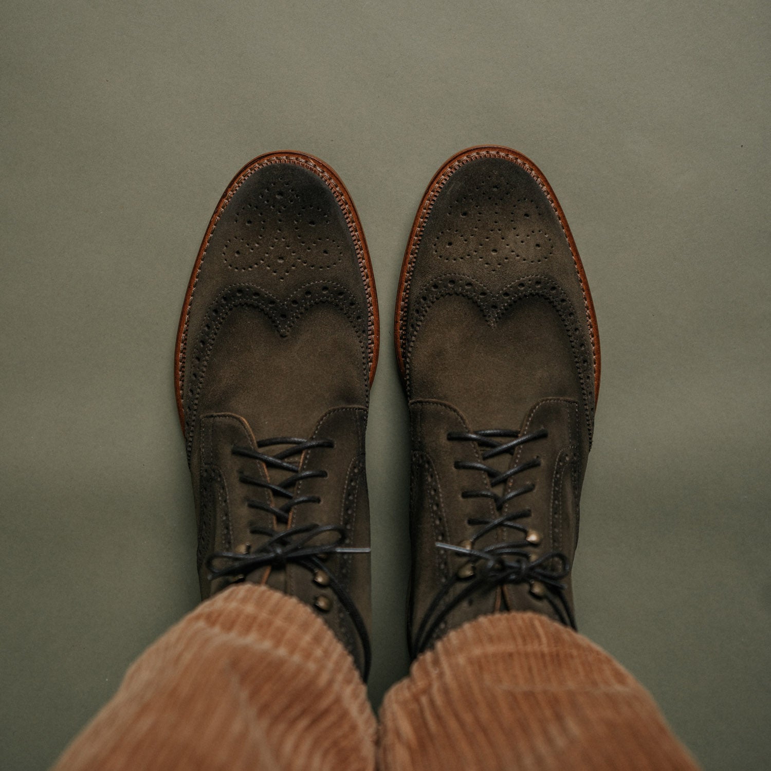 A top view of a person wearing brown suede lace-up dress shoes with perforated detailing and light brown corduroy pants against a green background.