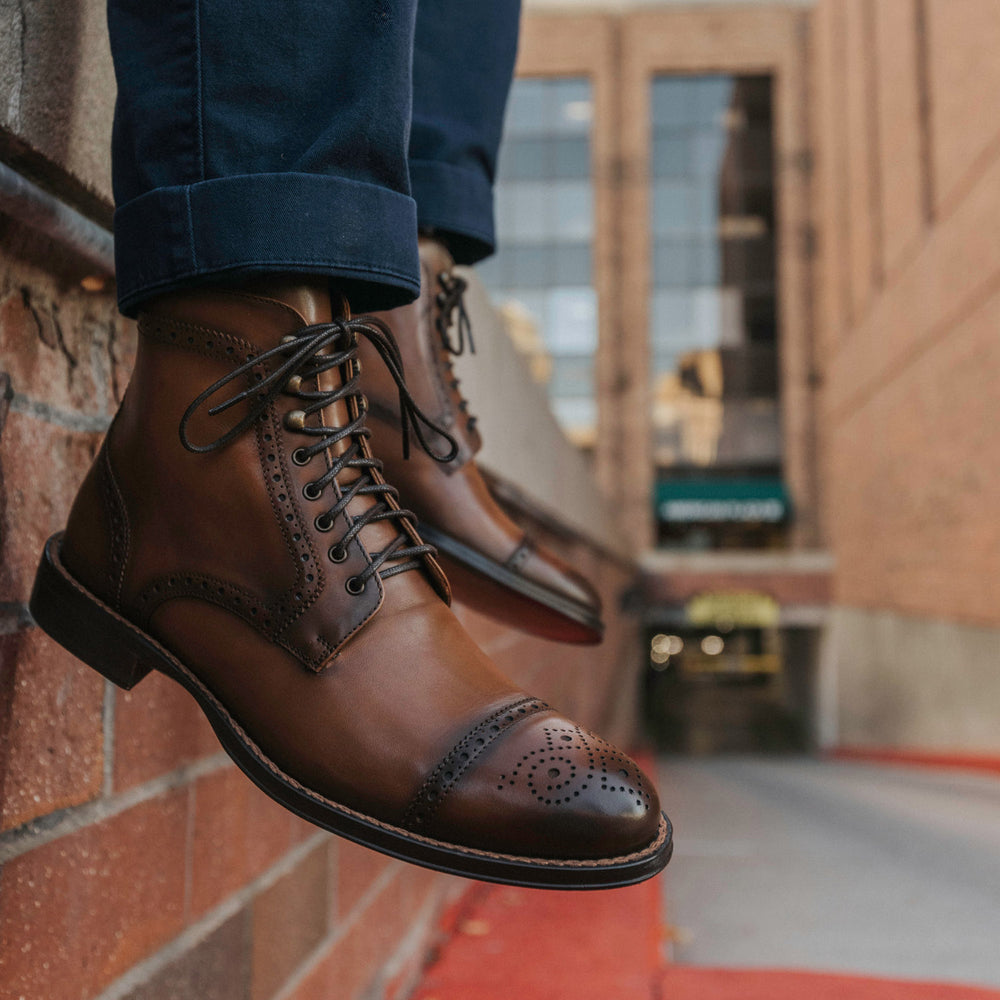 Close-up of a person wearing brown leather dress boots and dark blue jeans, standing near a brick wall with a blurred urban background.