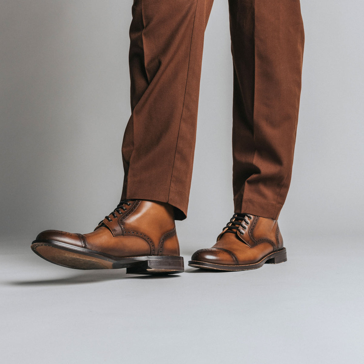 A person wearing brown leather lace-up shoes and brown trousers stands against a plain gray background.