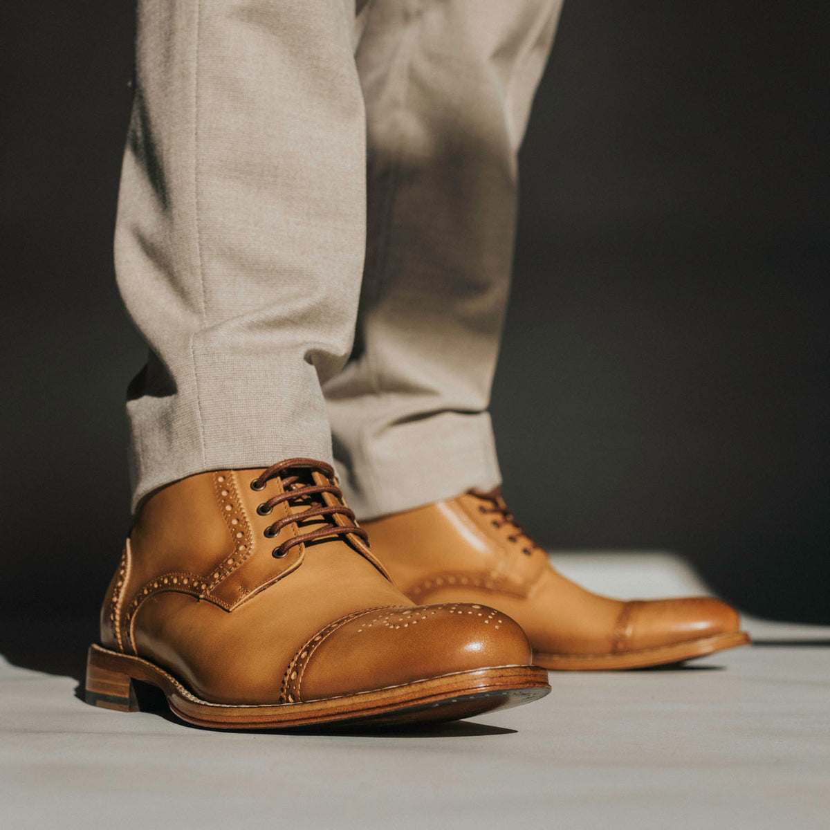 A person wearing tan leather dress shoes with brogue detailing and beige trousers stands on a surface with a dark background.