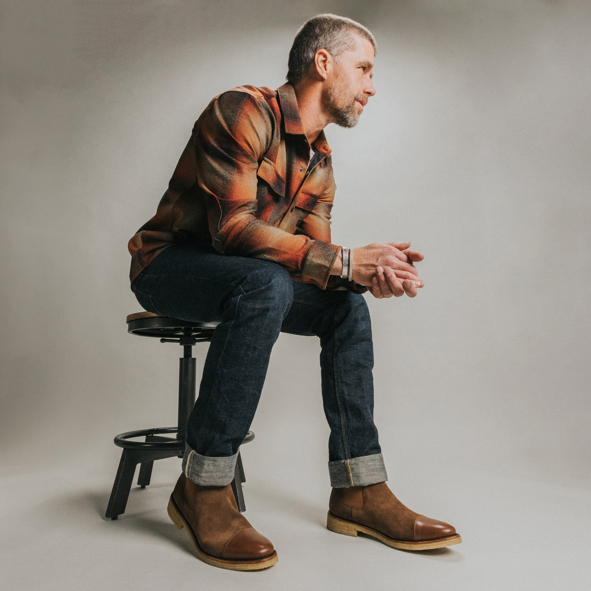 Man in a plaid shirt, jeans, and brown boots sits on a stool, facing sideways with hands clasped. Background is plain and neutral.