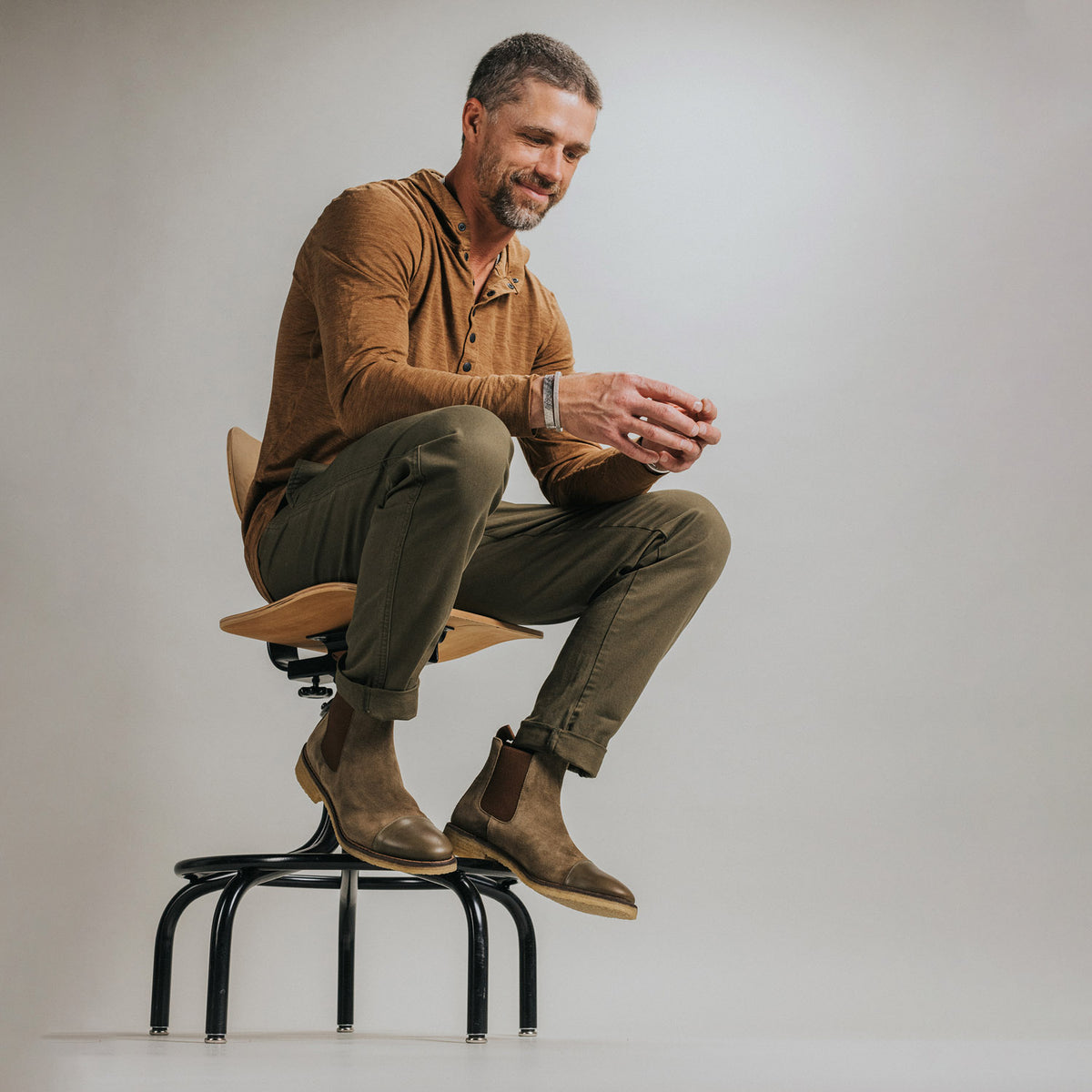 A man with short hair and a beard sits on a stool, dressed in a brown long-sleeve shirt, olive green pants, and brown boots, looking down while holding something in his hands.