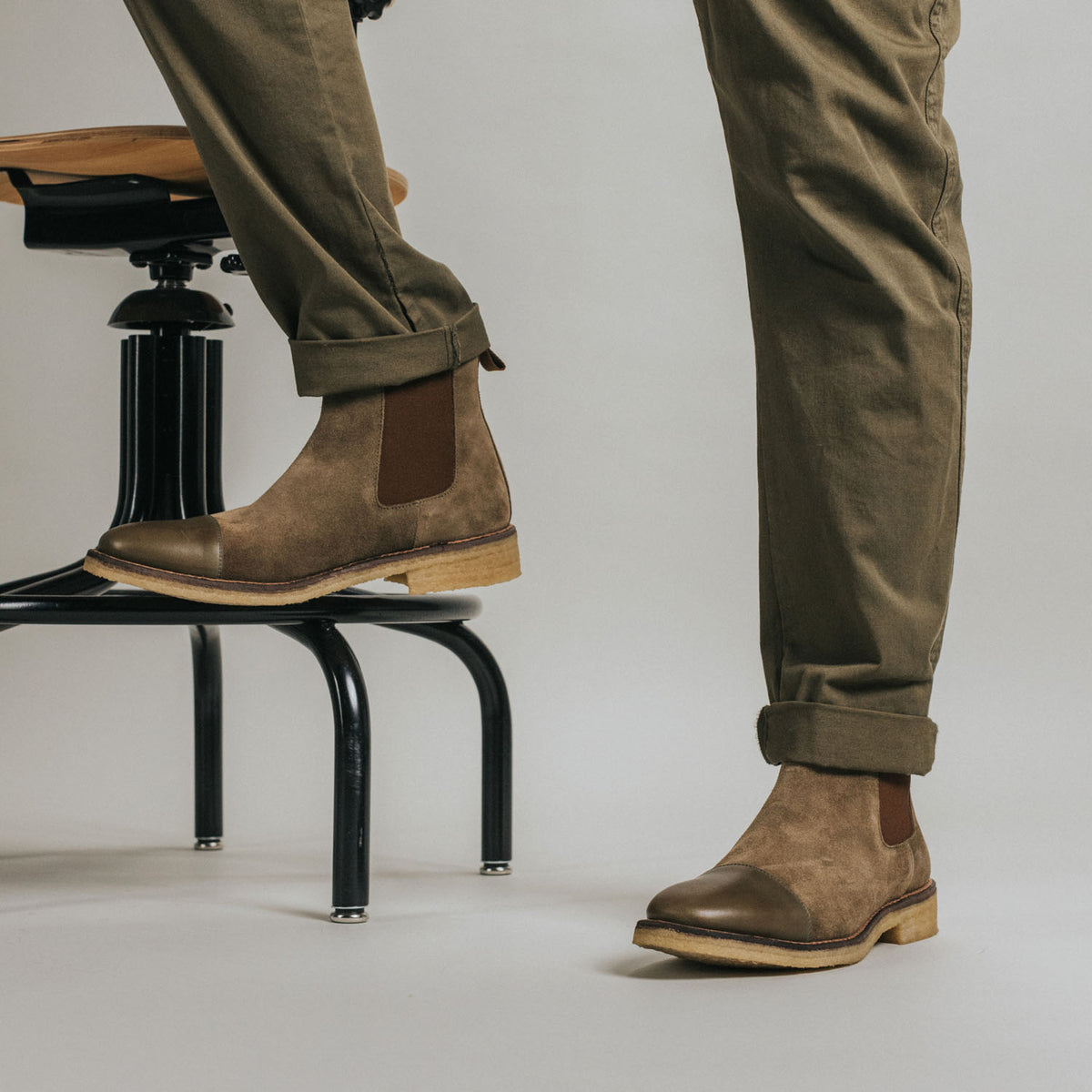 A person wearing light brown suede Chelsea boots with rolled-up khaki pants, standing near a backless swivel stool with black metal legs and a wooden seat.