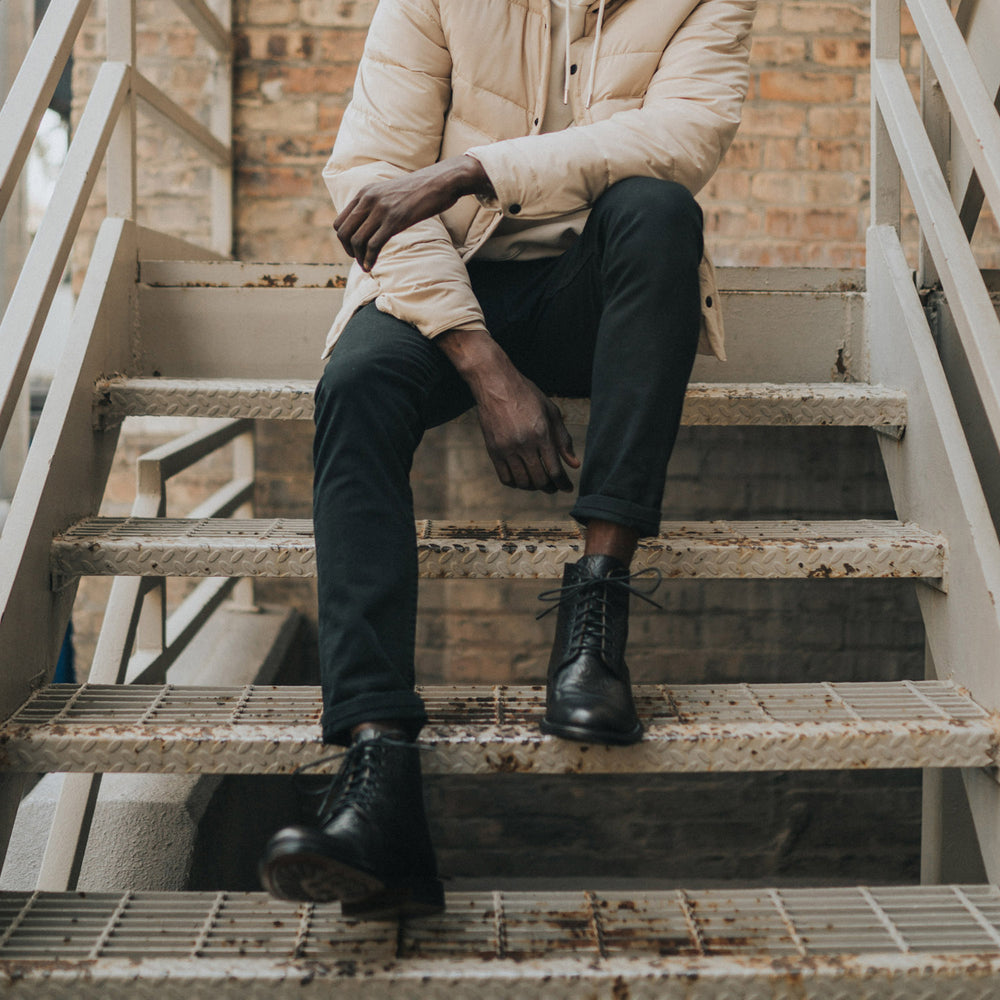 A person in a beige jacket and black pants sits on outdoor metal stairs, resting their arms on their knees.