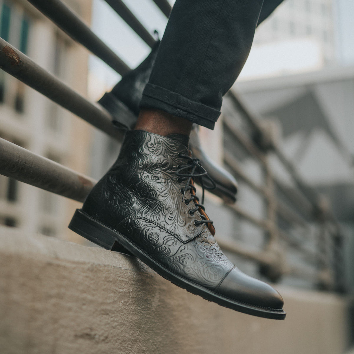 Close-up of a person wearing black dress shoes with intricate floral patterns, standing on a concrete ledge next to a metal railing.