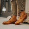 A person wearing brown leather boots with intricate designs stands on a sidewalk. The boots are paired with checkered pants.