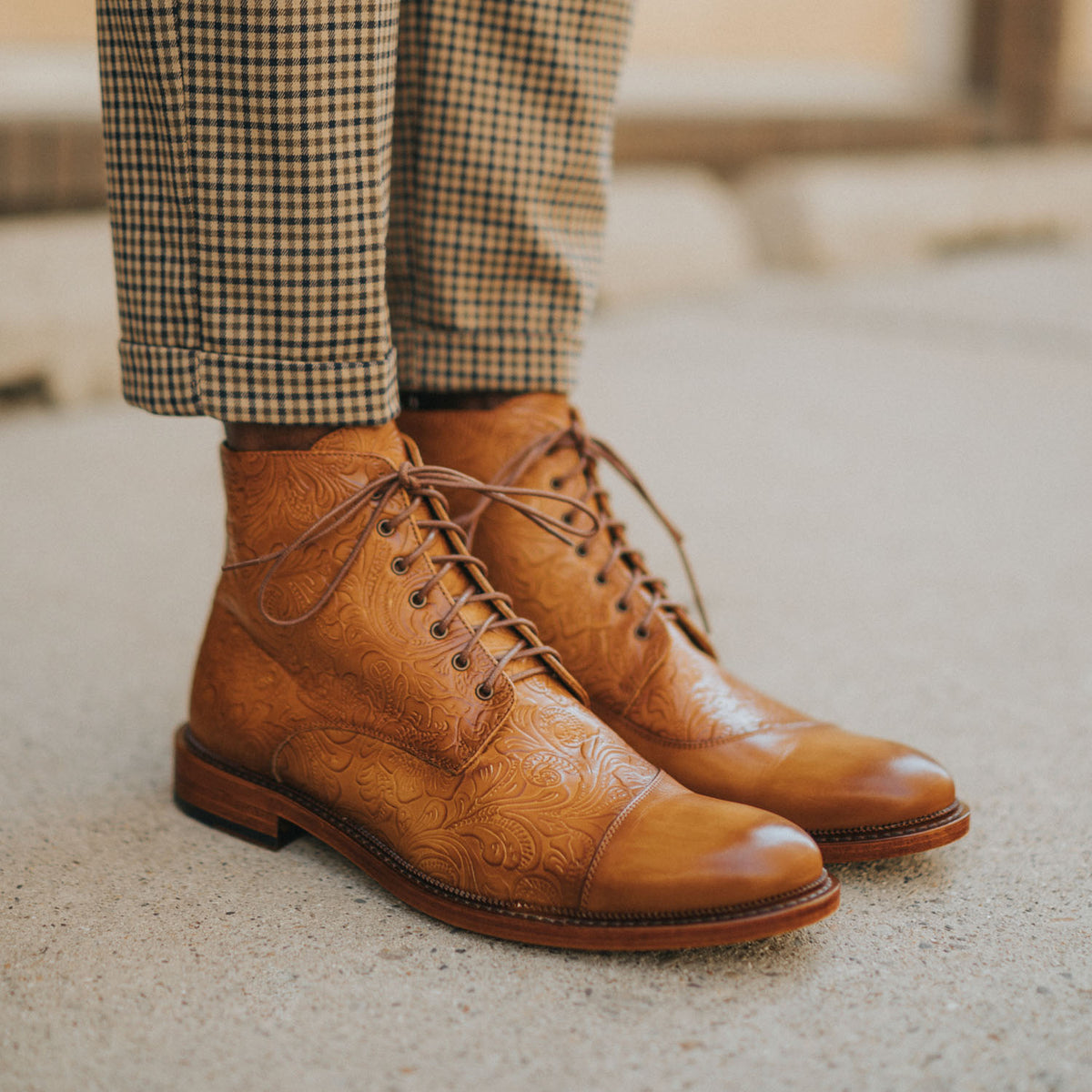 Close-up of a person wearing light brown, patterned leather boots and brown checkered pants, standing on a concrete surface.