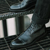 A person wearing black leather dress boots stands on a metal grate stairway.