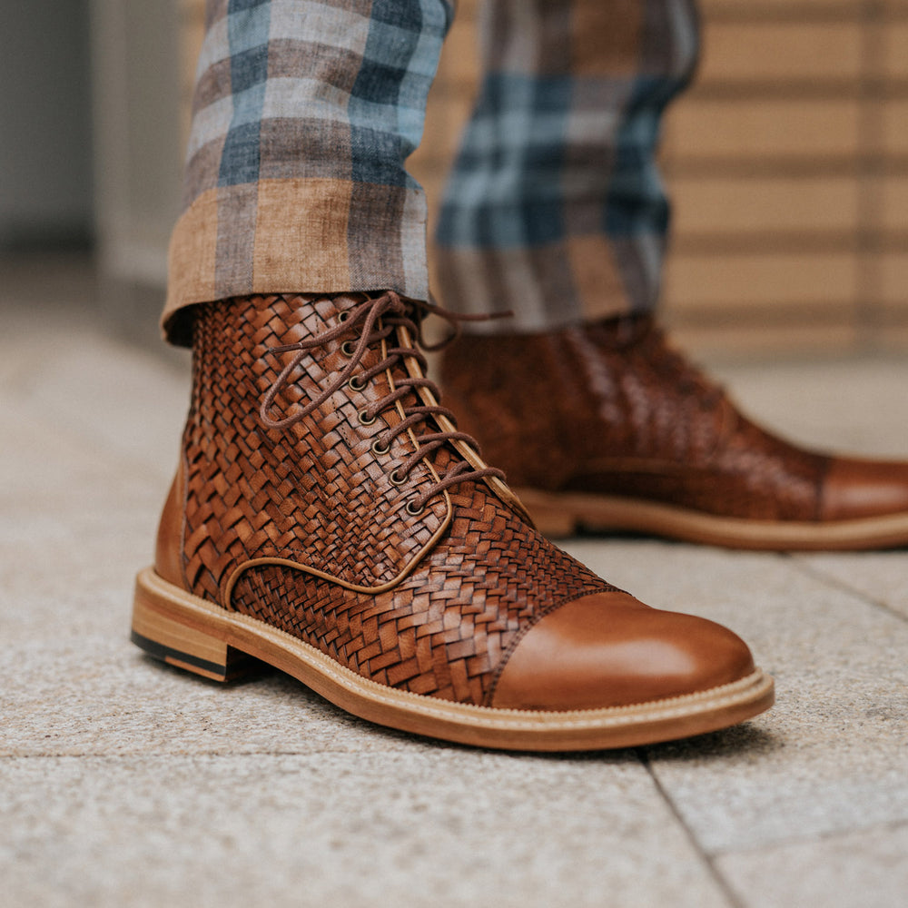 Person wearing brown woven leather boots and plaid pants stands on a tiled surface.