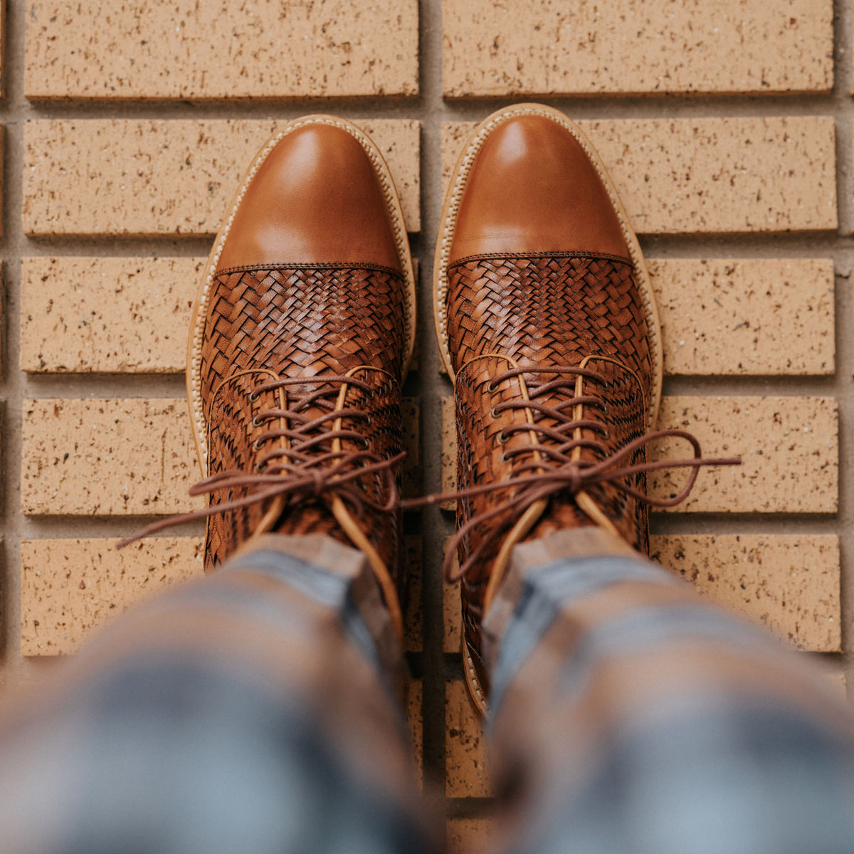 Top-down view of a person wearing brown woven leather boots, standing on a textured brick surface.