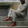 A man in a plaid suit and brown hat sits on concrete steps, showcasing brown leather boots.