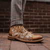 A person wearing checkered pants stands on a brick sidewalk, showcasing tan leather lace-up shoes with intricate floral embroidery.