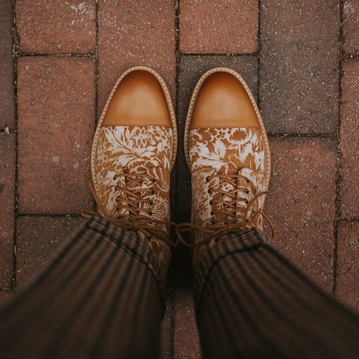 A person wearing patterned, brown leather shoes stands on a red brick pavement.