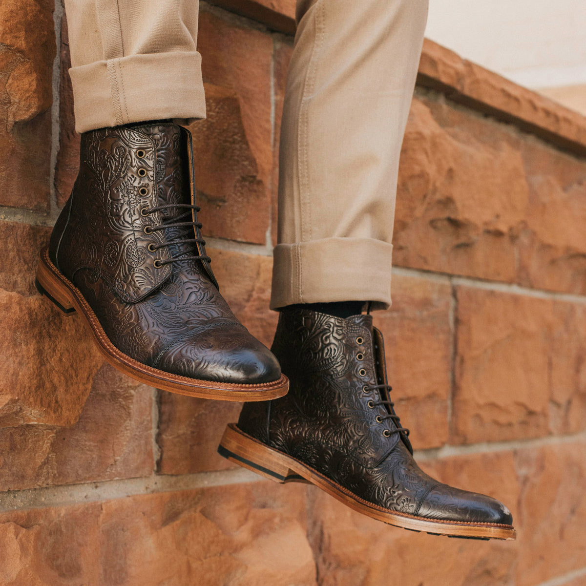 Person wearing intricately patterned dark leather boots and light khaki pants, standing against a brick wall.