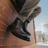 Person in plaid trousers sitting against a brick wall, showcasing brown leather boots with intricate designs.