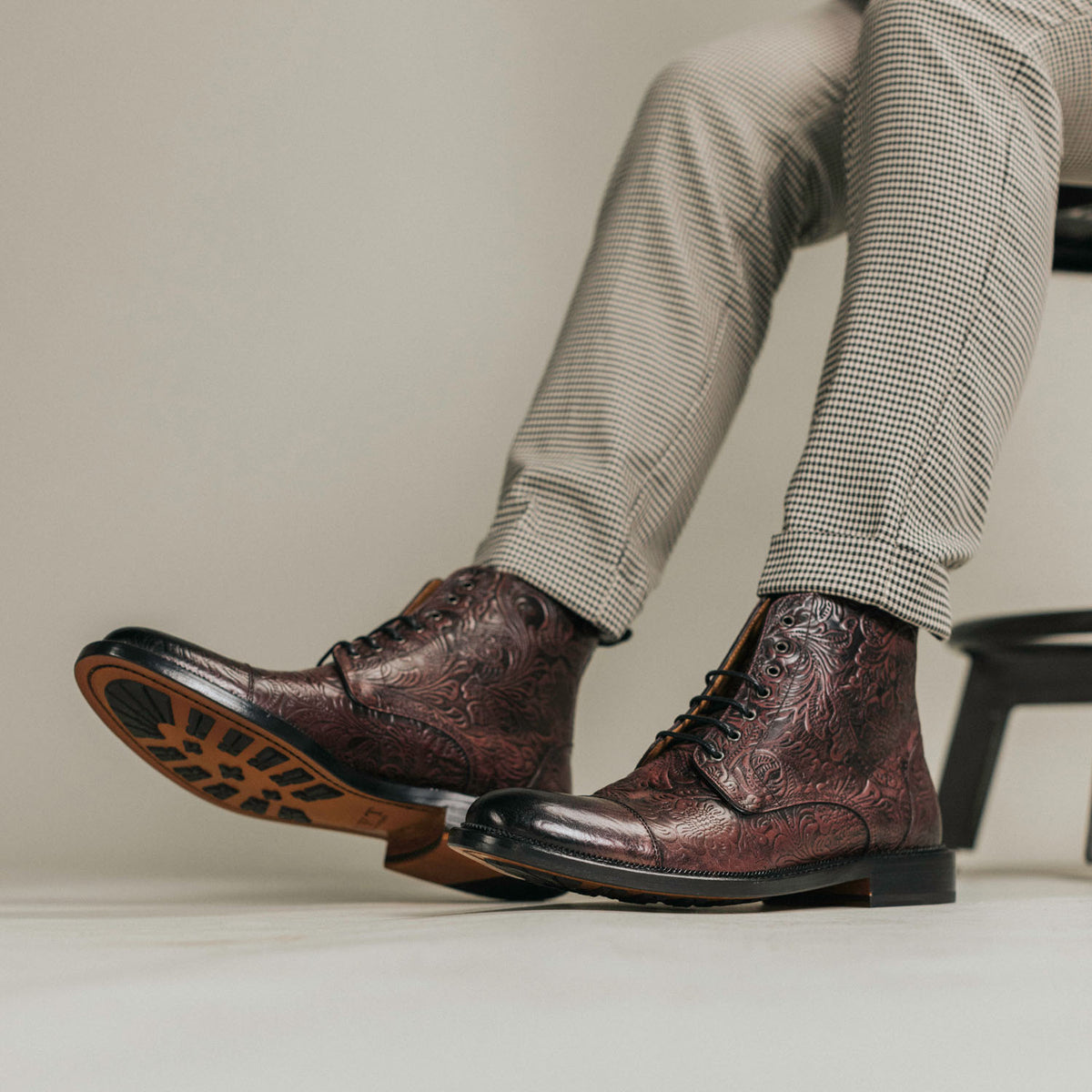 A person wearing patterned brown leather boots and checkered pants sits on a chair, with their legs crossed.